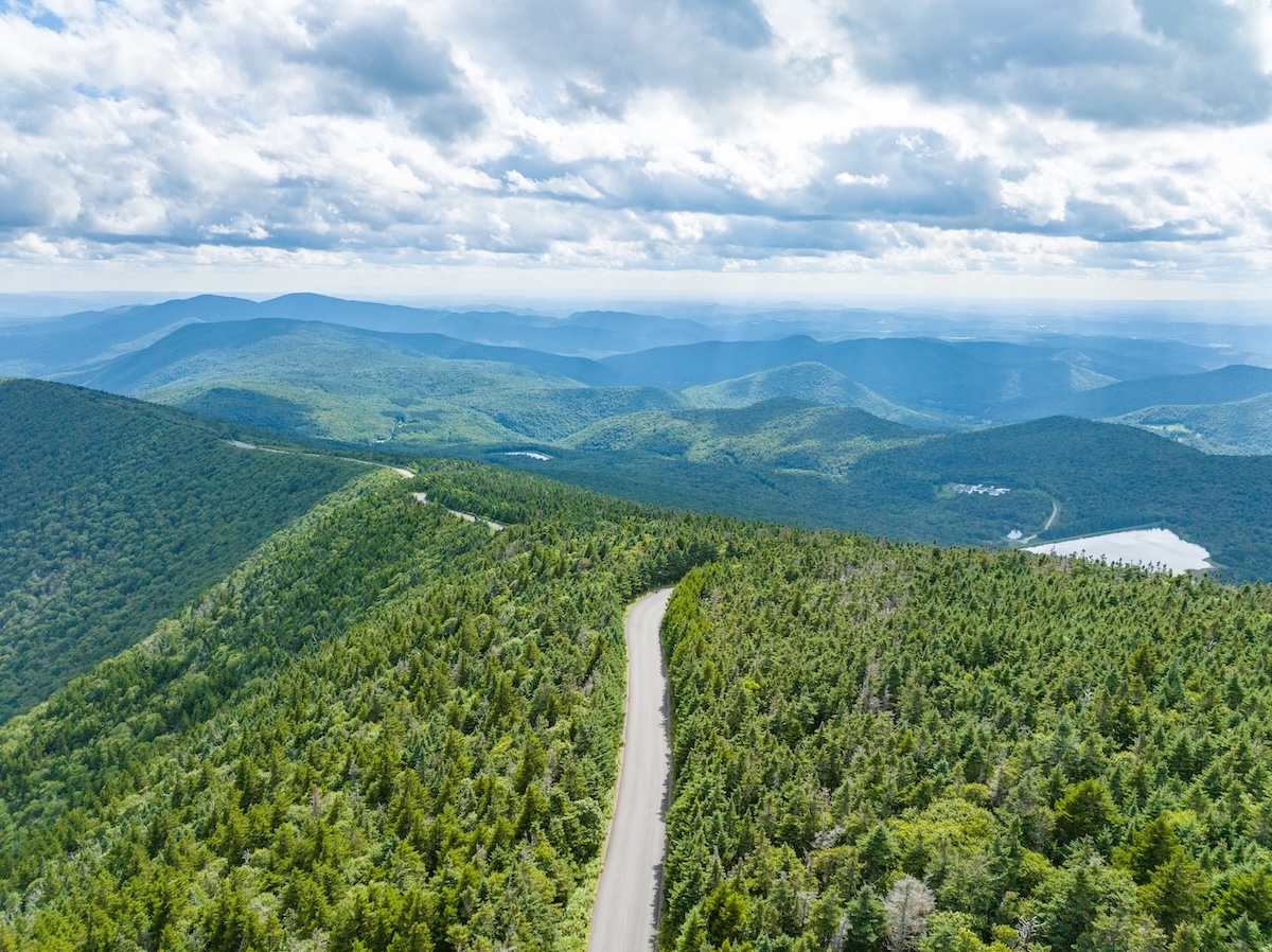 A winding road cuts through a dense green forest on a mountain, with rolling hills and a cloudy sky in the background.