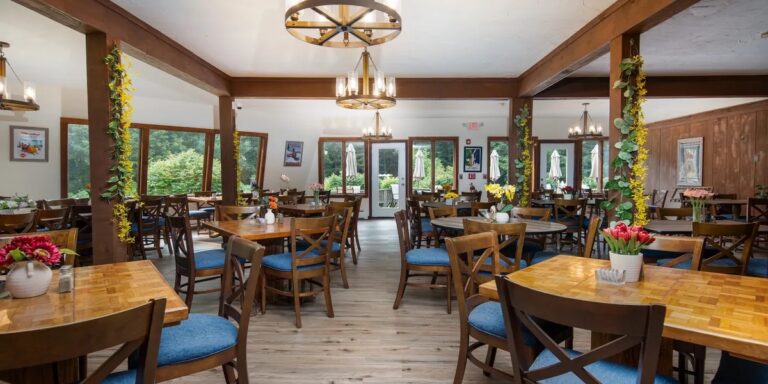 A spacious, well-lit dining room features wooden tables with blue-cushioned chairs, floral centerpieces, large windows, and decorative hanging lights.