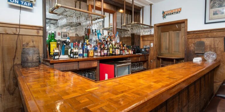 Wooden bar with a polished countertop, assorted liquor bottles, glassware hanging above, and a small fridge underneath. Wood paneling and signs decorate the walls.
