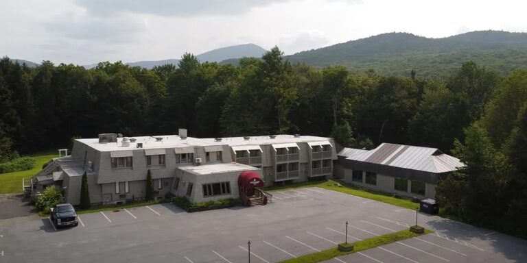Aerial view of a two-story building with a red awning, surrounded by trees and mountains, with an empty parking lot in front.