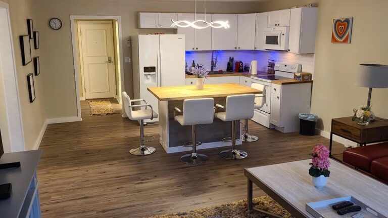 Modern kitchen with white cabinets, an island with three chairs, wood flooring, and blue under-cabinet lighting. Living area with coffee table and pink flowers visible in foreground.