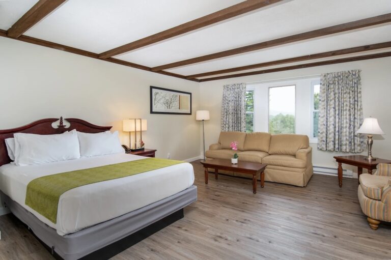 A hotel room with a king bed, beige sofa, armchair, two coffee tables, lamps, wall art, and large window with floral curtains, featuring wood floors and ceiling beams.
