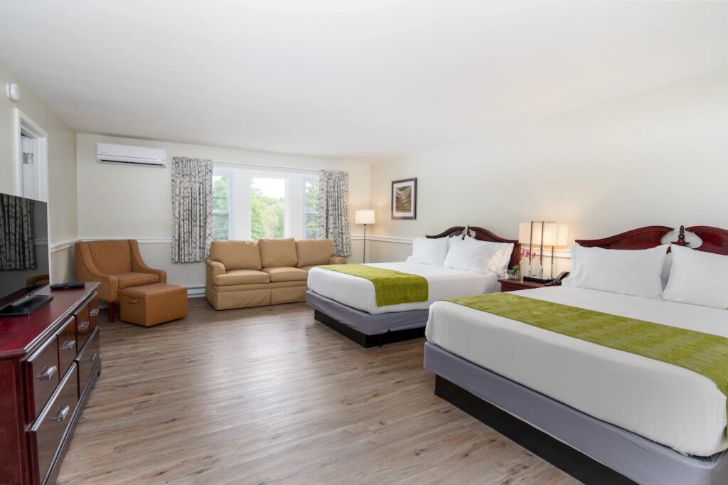 A hotel room with two double beds, a sofa, armchair, TV, and a window with curtains; the room has light walls and wooden flooring.