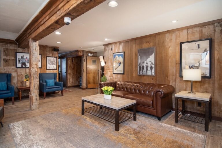 A cozy lounge with a brown leather sofa, marble coffee table, blue armchairs, framed art on wood-paneled walls, and a table lamp on a side table.