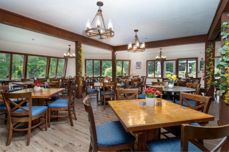 A spacious dining room with wooden tables and chairs, blue seat cushions, large windows, floral decorations, and chandeliers hanging from the ceiling.