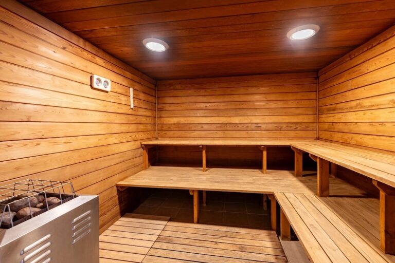 Interior of a wooden sauna room with benches along the walls, a metal stove in the corner, and two ceiling lights.