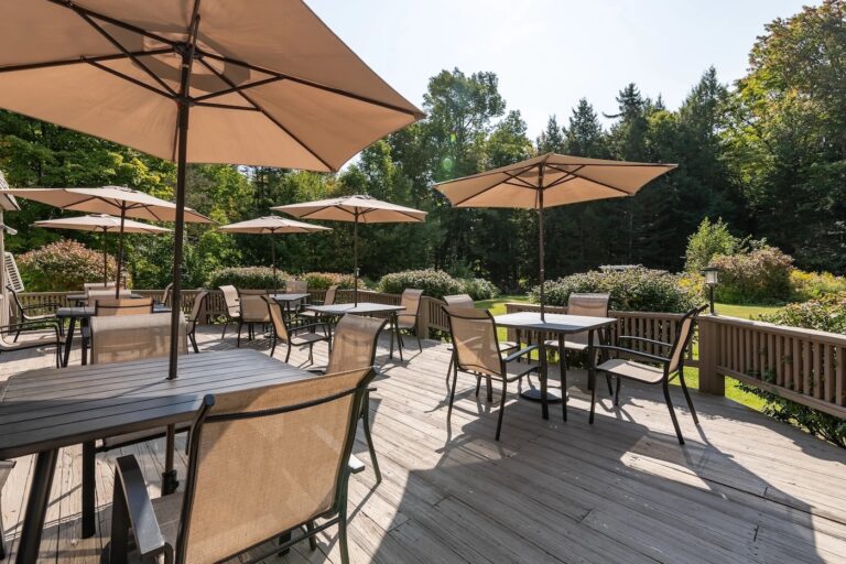 Outdoor patio with empty tables and chairs under large beige umbrellas, surrounded by greenery and trees on a sunny day.