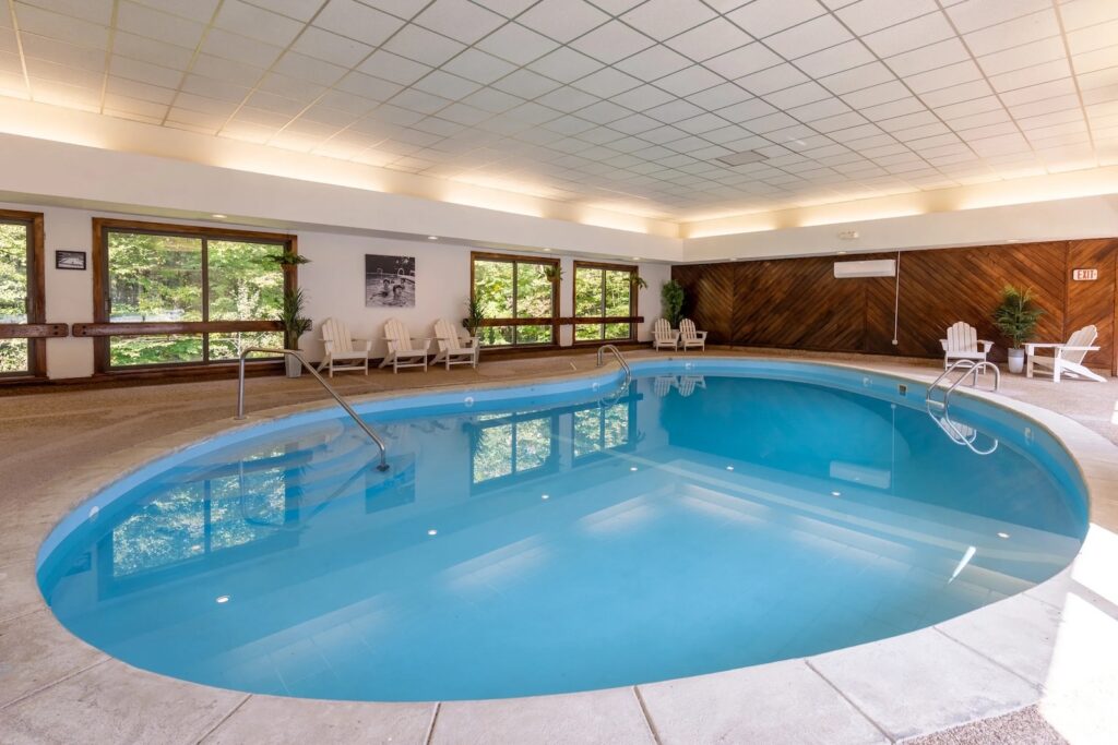 An indoor swimming pool with blue water, surrounded by white lounge chairs, large windows with outdoor views, and a wood-paneled wall.