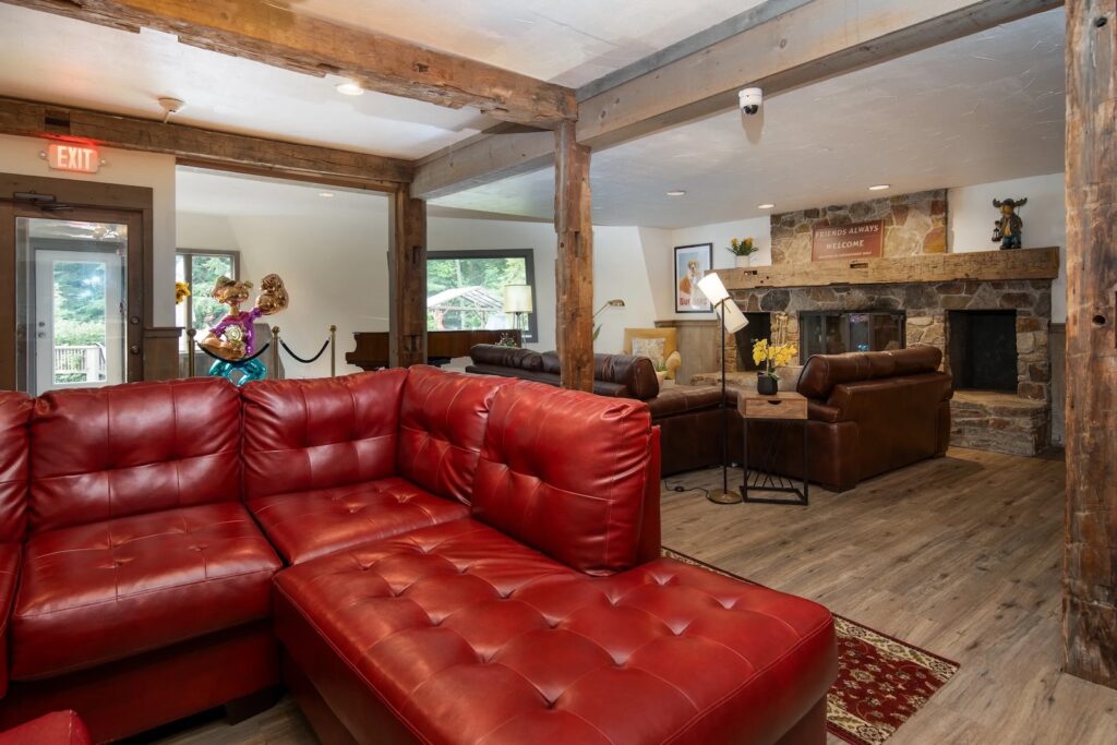 A rustic lounge area with exposed wooden beams, red leather sectional sofa, brown leather armchairs, stone fireplace, wooden floors, and natural light from windows.