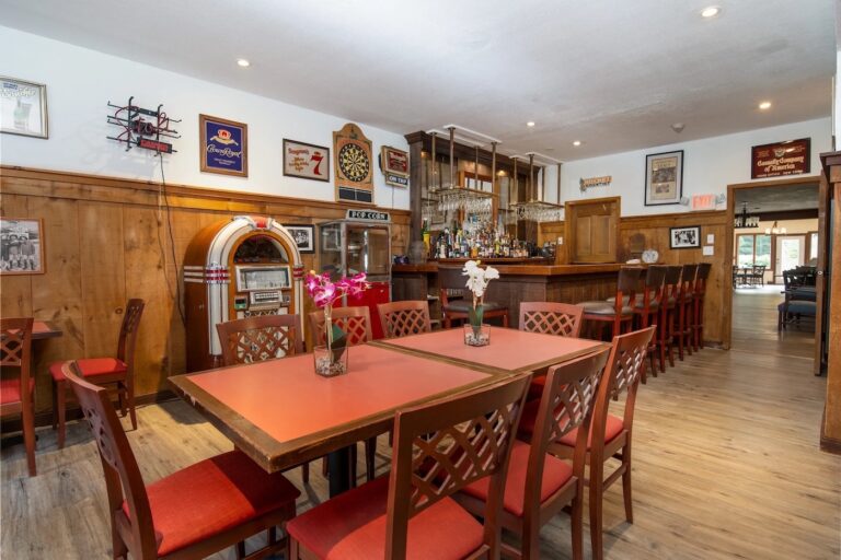A bar area with wooden tables and red chairs, a jukebox, a dartboard, wall art, and a well-stocked bar in a cozy, warmly lit setting.