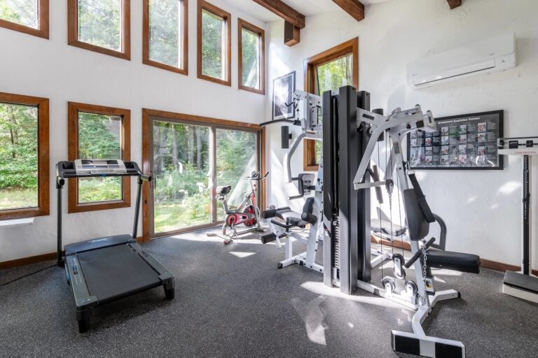 Home gym with treadmill, multi-station weight machine, and exercise bike. Large windows provide natural light and a view of greenery outside.
