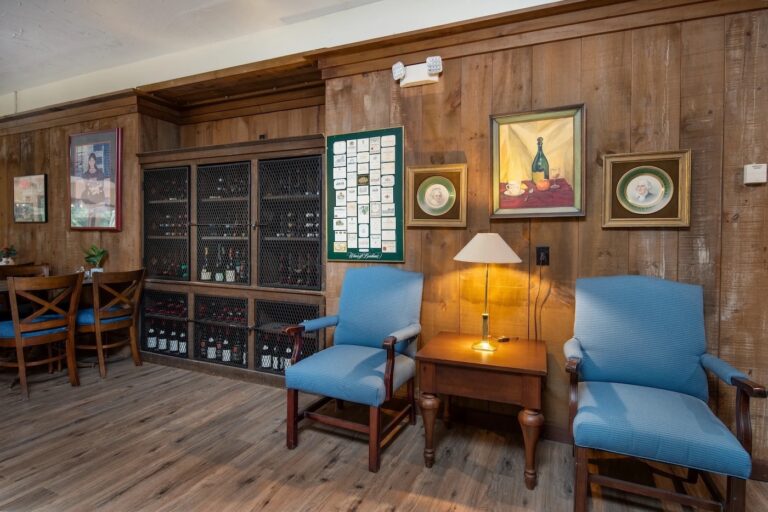 Two blue chairs and a wooden side table with a lamp sit against a wood-paneled wall, with framed art, a wine cabinet, and decorative plates in the background.