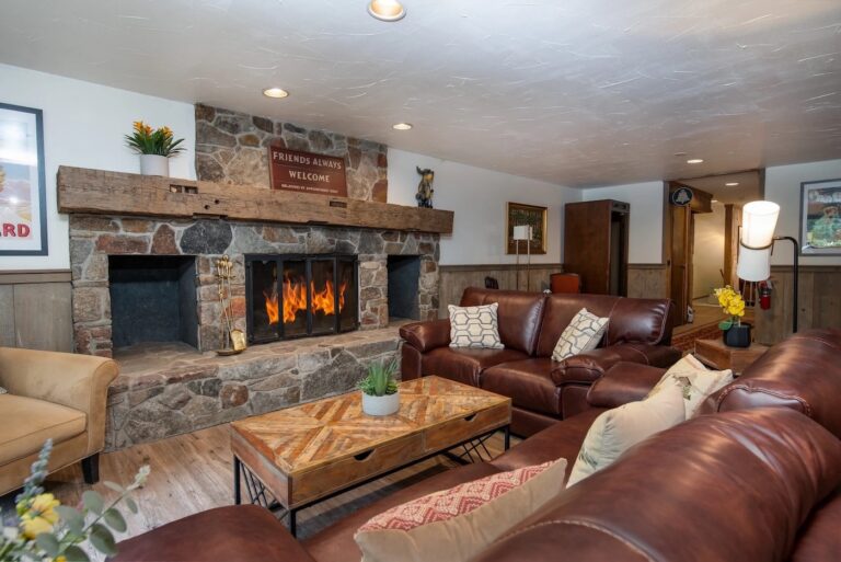 A cozy living room with brown leather sofas, a stone fireplace with a fire burning, wooden coffee table, and wall art above the mantel.