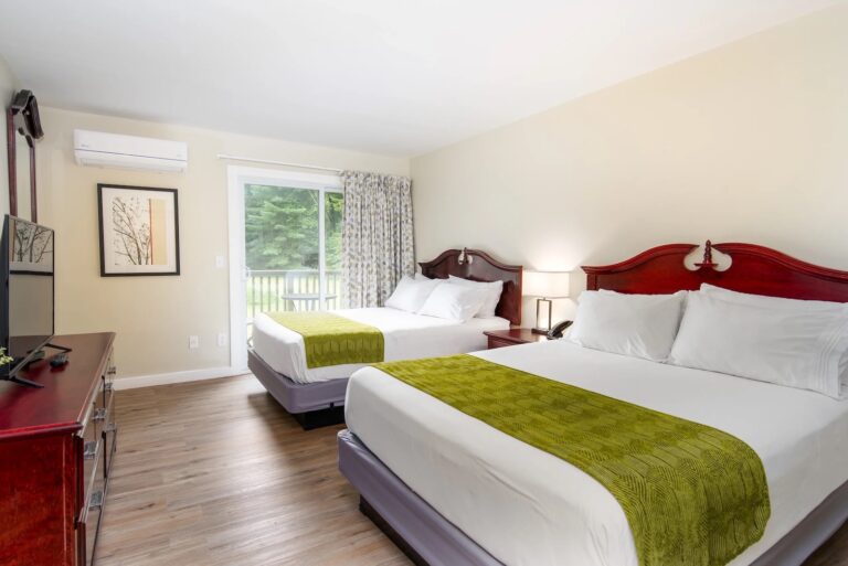 A hotel room with two double beds, white linens, green throws, a TV, dresser, lamp, artwork, and a sliding glass door leading outside.