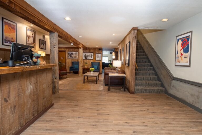 A rustic hotel lobby with a wooden front desk, seating area with chairs and tables, framed art on the walls, and a carpeted staircase on the right.
