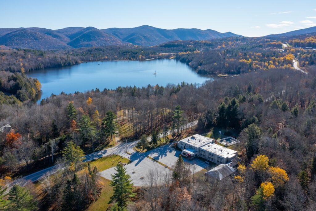 Aerial view of a lake surrounded by autumn trees and hills, with a building and parking lot in the foreground under a clear blue sky.