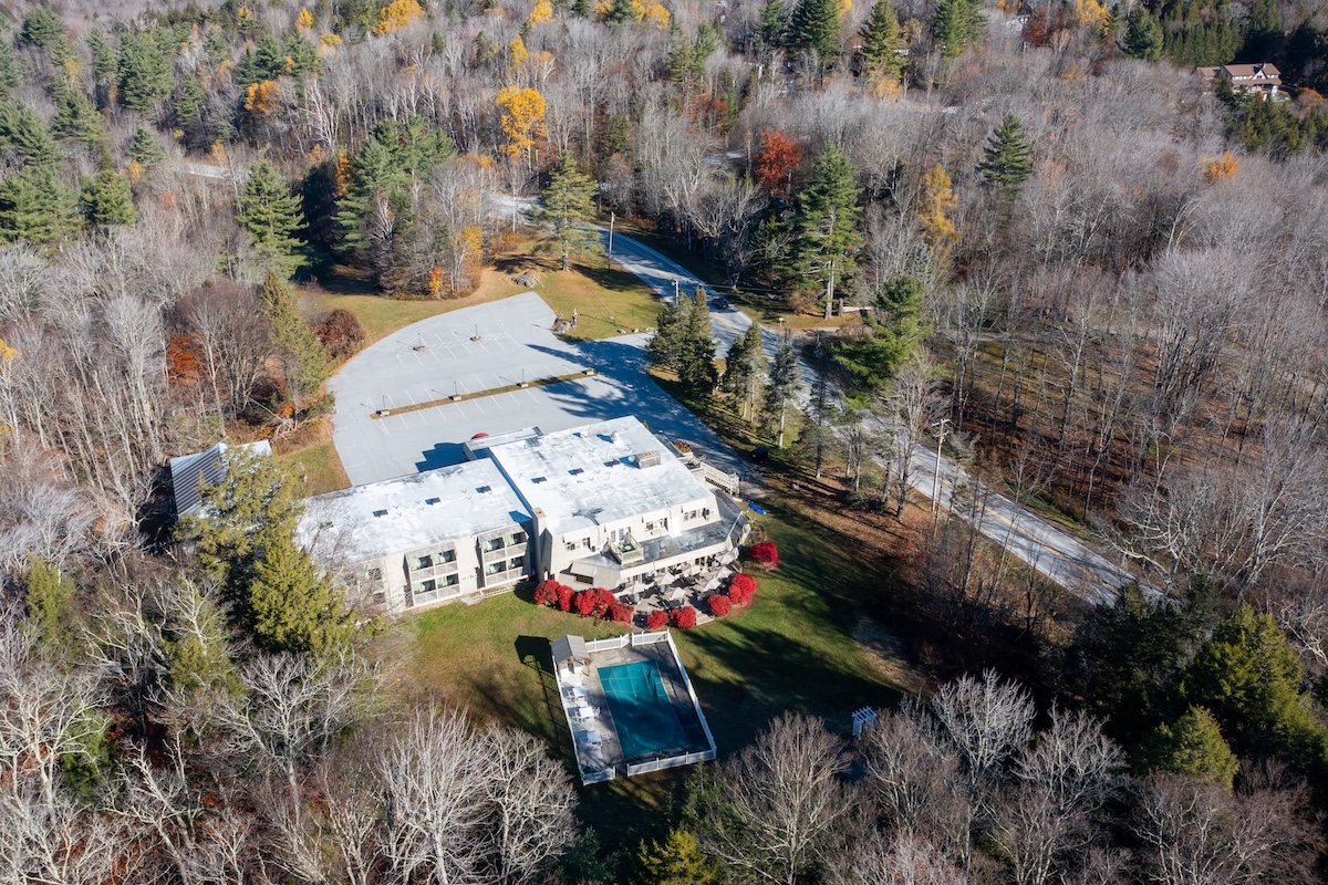 Aerial view of a large, modern house with a swimming pool, surrounded by trees with autumn foliage and a parking lot nearby.