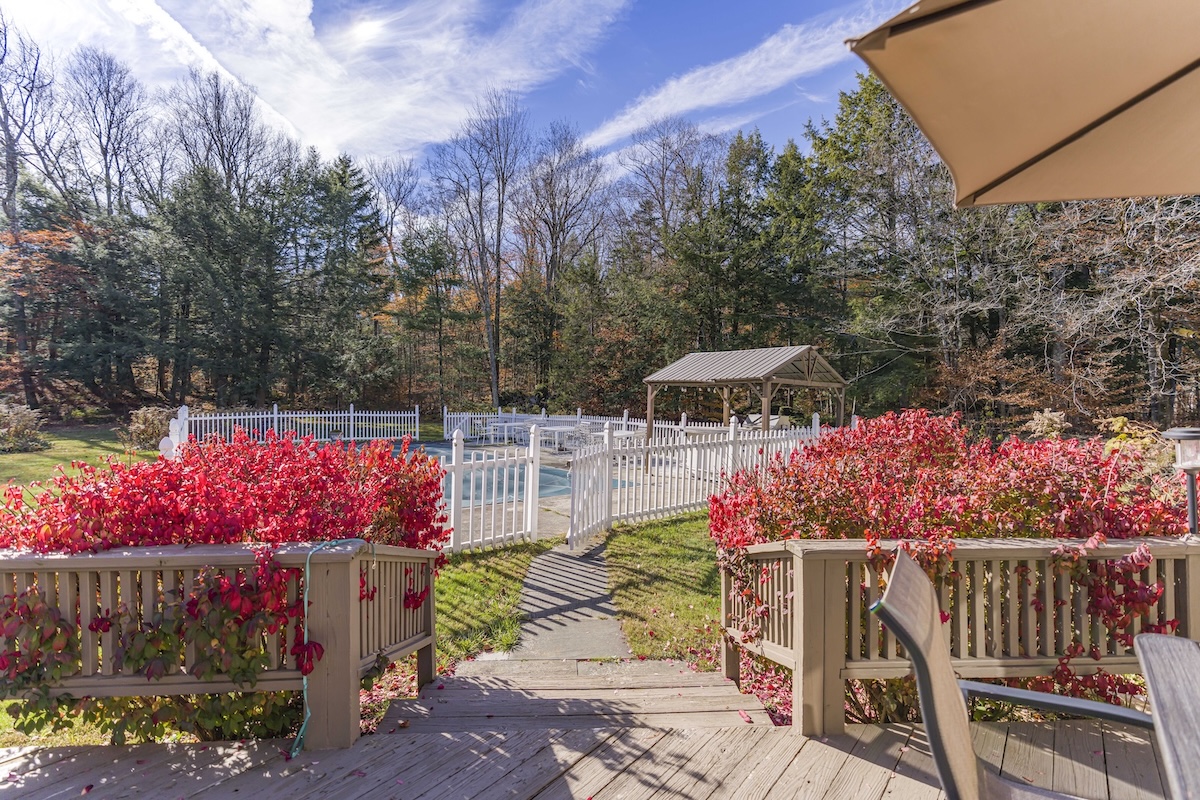 View from a wooden deck with red foliage, leading to a fenced outdoor pool and a gazebo, with trees and a blue sky in the background.
