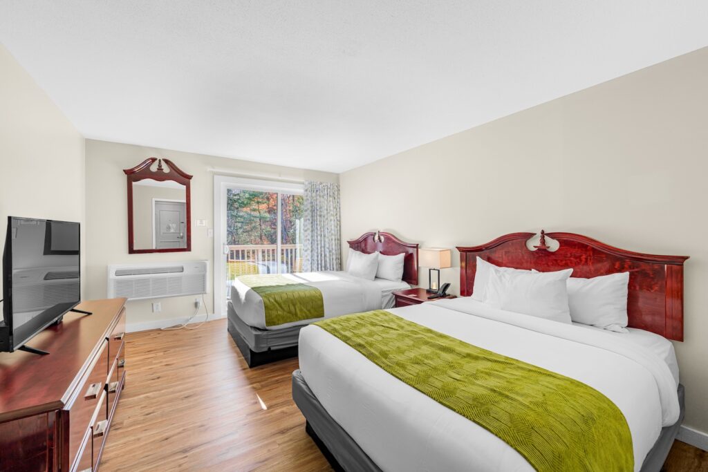 Hotel room with two double beds, wooden headboards, a TV on a dresser, a mirror, and a window with natural light and a view of trees outside.