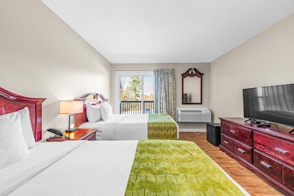 A hotel room with two beds, green bed runners, a wooden dresser, TV, mirror, air conditioner, lamp, and a window with curtains showing trees outside.