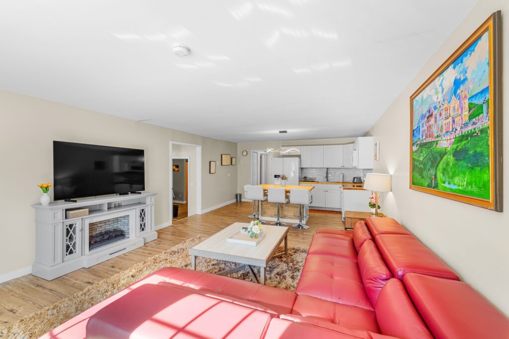 Bright living room with a large red sectional sofa, wall-mounted TV, electric fireplace, and open kitchen with white cabinets and bar stools. Colorful painting hangs on the wall.
