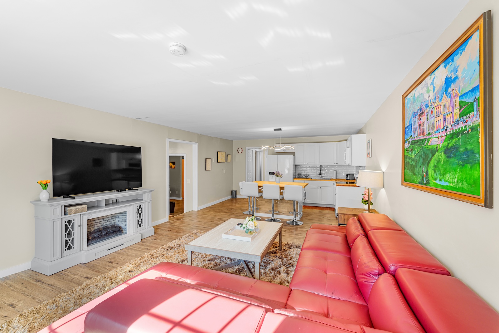 Bright living room with a large red sectional sofa, wall-mounted TV, electric fireplace, and open kitchen with white cabinets and bar stools. Colorful painting hangs on the wall.