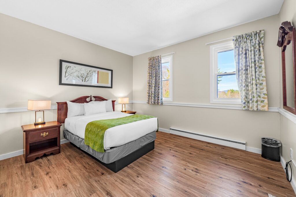 A neatly made bed with green blanket in a simple, bright bedroom with wood flooring, two windows with curtains, two bedside tables, and a large wall mirror.