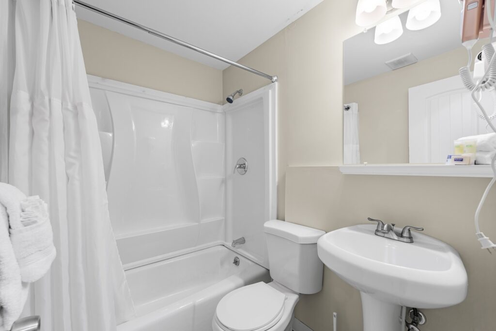 A small bathroom with a white bathtub and shower, a toilet, a pedestal sink, a wall-mounted mirror, and beige walls under bright lighting.