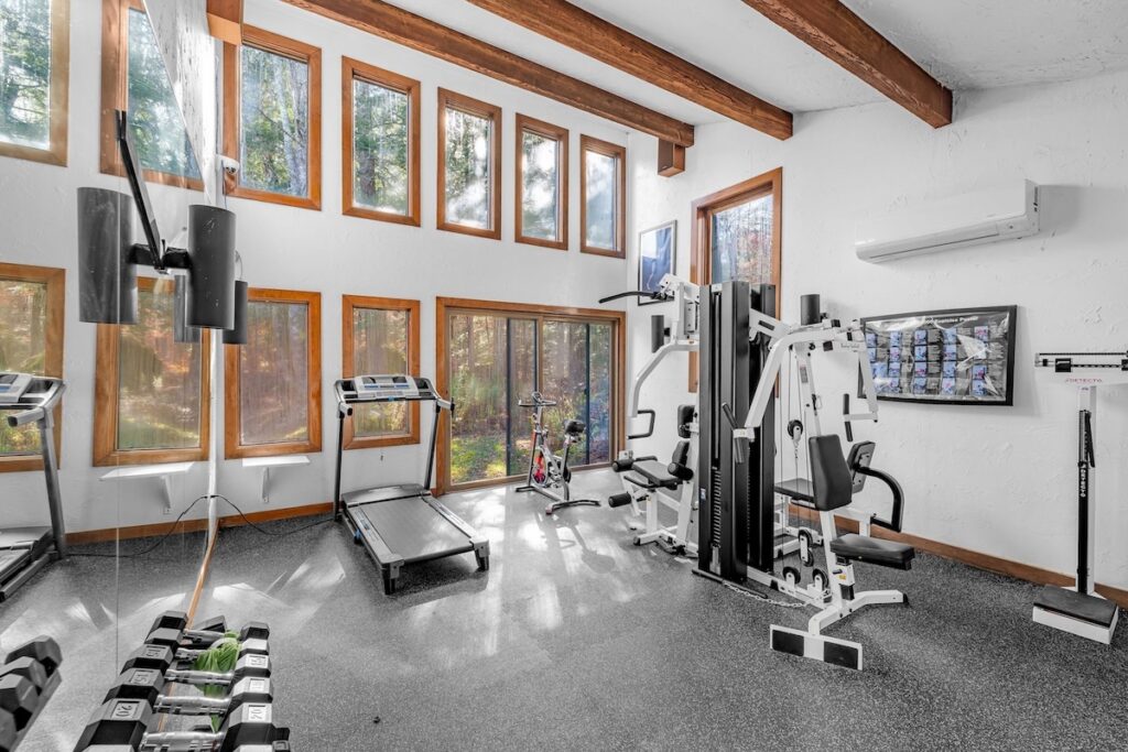 A home gym with large windows, featuring a treadmill, stationary bike, multi-use weight machine, bench, wall-mounted TV, and a rack of dumbbells.