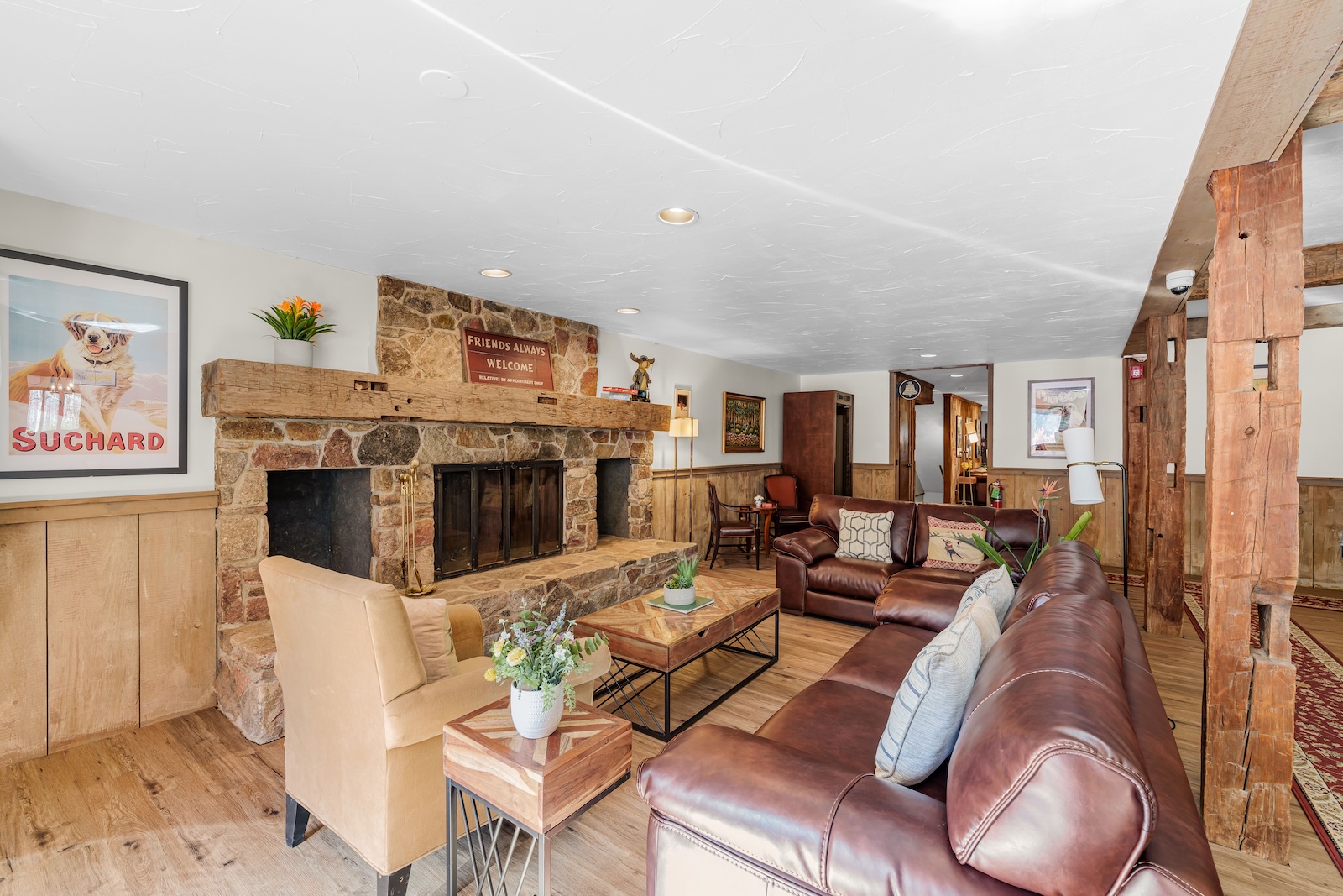 Cozy living room with brown leather sofas, wooden tables, a stone fireplace, wall art, and wood-paneled walls in a rustic interior setting.