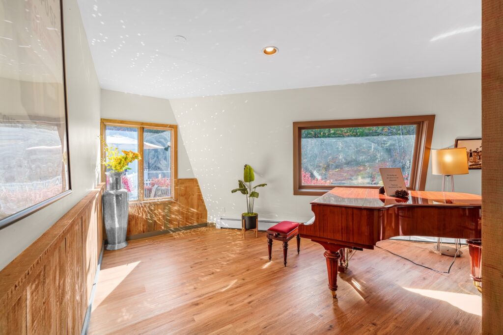 A bright room with wood floors features a grand piano, a red bench, a potted plant, a large vase with yellow flowers, and big windows letting in natural light.