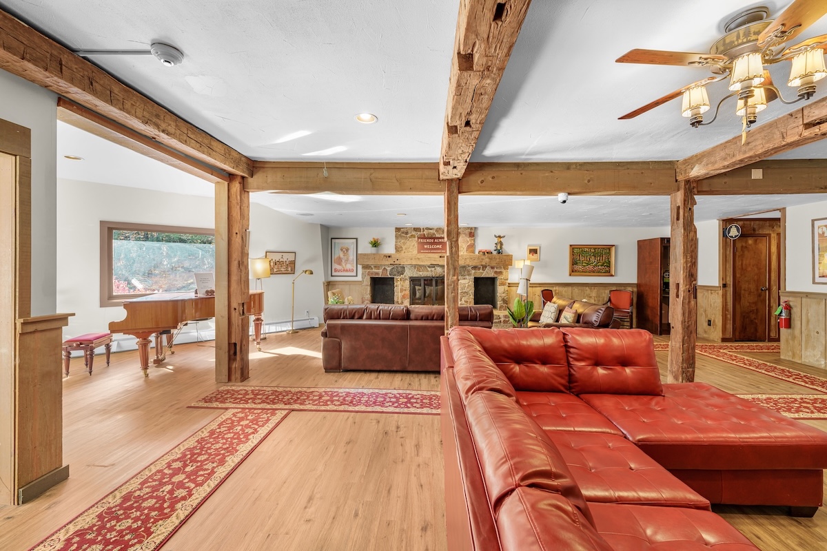 Spacious living room with exposed wooden beams, red sectional sofa, stone fireplace, grand piano, and wooden floor with patterned rugs.