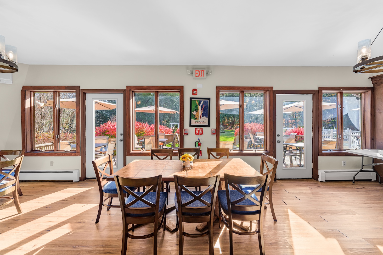 A dining area with a wooden table and chairs, large windows, and glass doors overlooking a patio with umbrellas and autumn foliage.