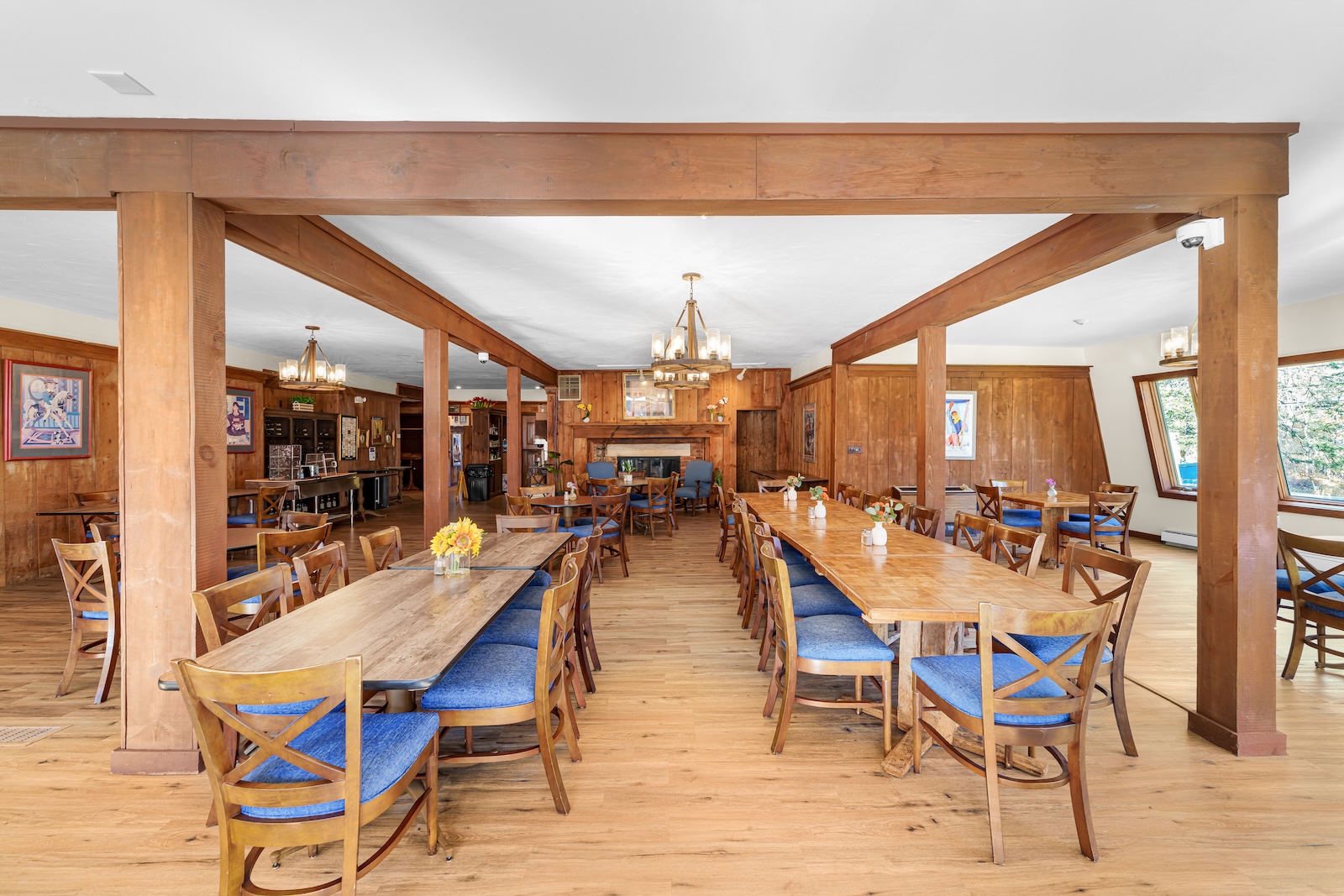 A spacious dining room with wooden floors, several wooden tables and chairs with blue cushions, chandeliers, and wall art.