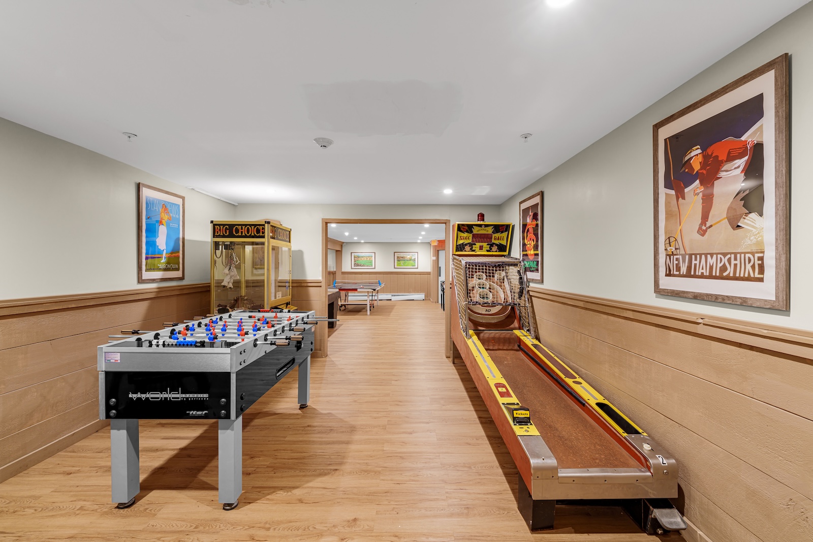 A game room with a foosball table, a skee-ball machine, a ping pong table in the background, and vintage-style posters on the walls.