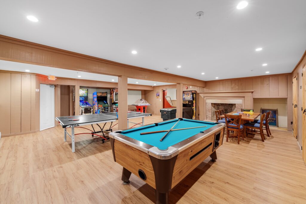 A basement game room with a pool table, air hockey table, foosball, arcade games, wooden chairs and tables, and wood-paneled walls and flooring.