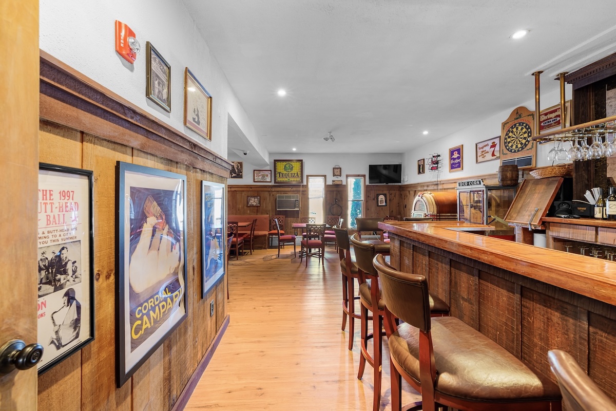 Bar interior with wooden flooring and walls, high chairs along a counter, wall art, a dartboard, tables, and a television in the background.